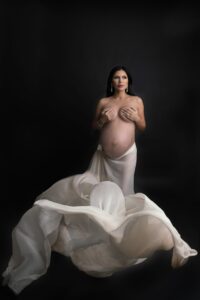 Creative Maternity Accessories. A pregnant woman in a white dress posing on a black background.
