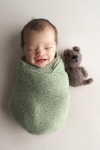 A newborn wrapped in a green wrap with a teddy bear and smiles.