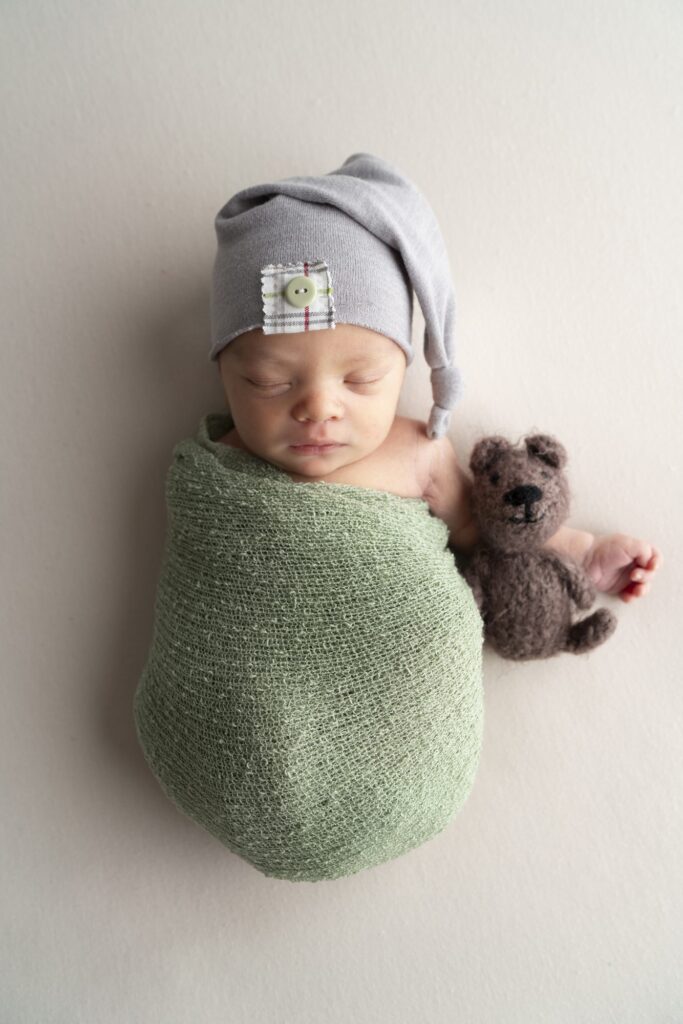 A newborn wrapped in a green blanket with a teddy bear and smiles.