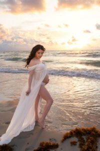 A pregnant woman in a flowing white dress stands barefoot on the beach at sunset, gentle waves and clouds behind her—captured beautifully by a Miami maternity photographer.