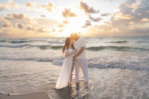 A couple stands close together on a beach at sunset, dressed in white, as waves roll in behind them and sunlight illuminates their embrace—captured beautifully by a Miami maternity photographer.
