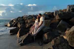 A couple dressed in white sits on large rocks by the ocean at sunset, with waves approaching and clouds in the sky—an idyllic moment beautifully captured by a Miami maternity photographer.