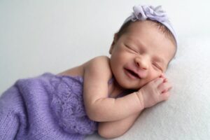 Newborn baby sleeping on a white surface, wrapped in a purple blanket, wearing a matching headband, and smiling with eyes closed.