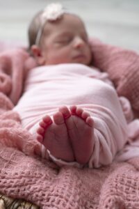 A newborn baby wrapped in a pink blanket sleeps with feet in focus, lying on a textured pink fabric.