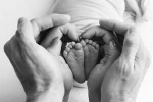 Close-up of adult hands forming a heart shape around a newborn baby's feet, with the baby lying on a soft surface.