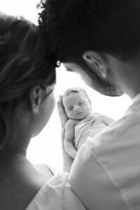 A man and woman gently cradle a sleeping newborn baby wrapped in a blanket, viewed from behind the adults in soft lighting.