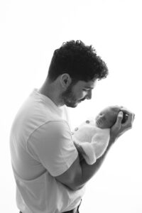 A man in a white shirt holds a sleeping newborn baby wrapped in a knitted blanket against a plain white background.