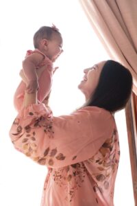 A woman in a floral robe holds a smiling baby up near a window with light streaming in, capturing a heartfelt moment perfect for a Miami family photographer.