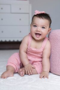 A baby wearing a pink outfit and hair bow sits on a light carpeted floor, smiling with an open mouth next to a round pink cushion—captured beautifully by a Miami family photographer.
