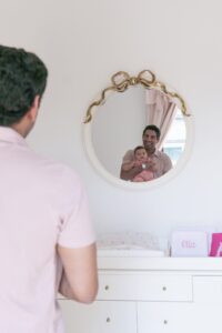 A man holding a baby is seen in the reflection of a round, gold-rimmed mirror above a white dresser with baby items and a name sign reading "Ella," perfectly captured by a Miami family photographer.