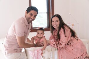 A man and woman smile beside a baby standing in a crib in a brightly lit nursery decorated with pink and white accents, captured beautifully by a Miami family photographer.