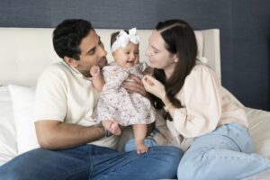 A man and woman sit on a bed holding a smiling baby dressed in a floral outfit and headband, all appearing happy—captured beautifully by a Miami family photographer.