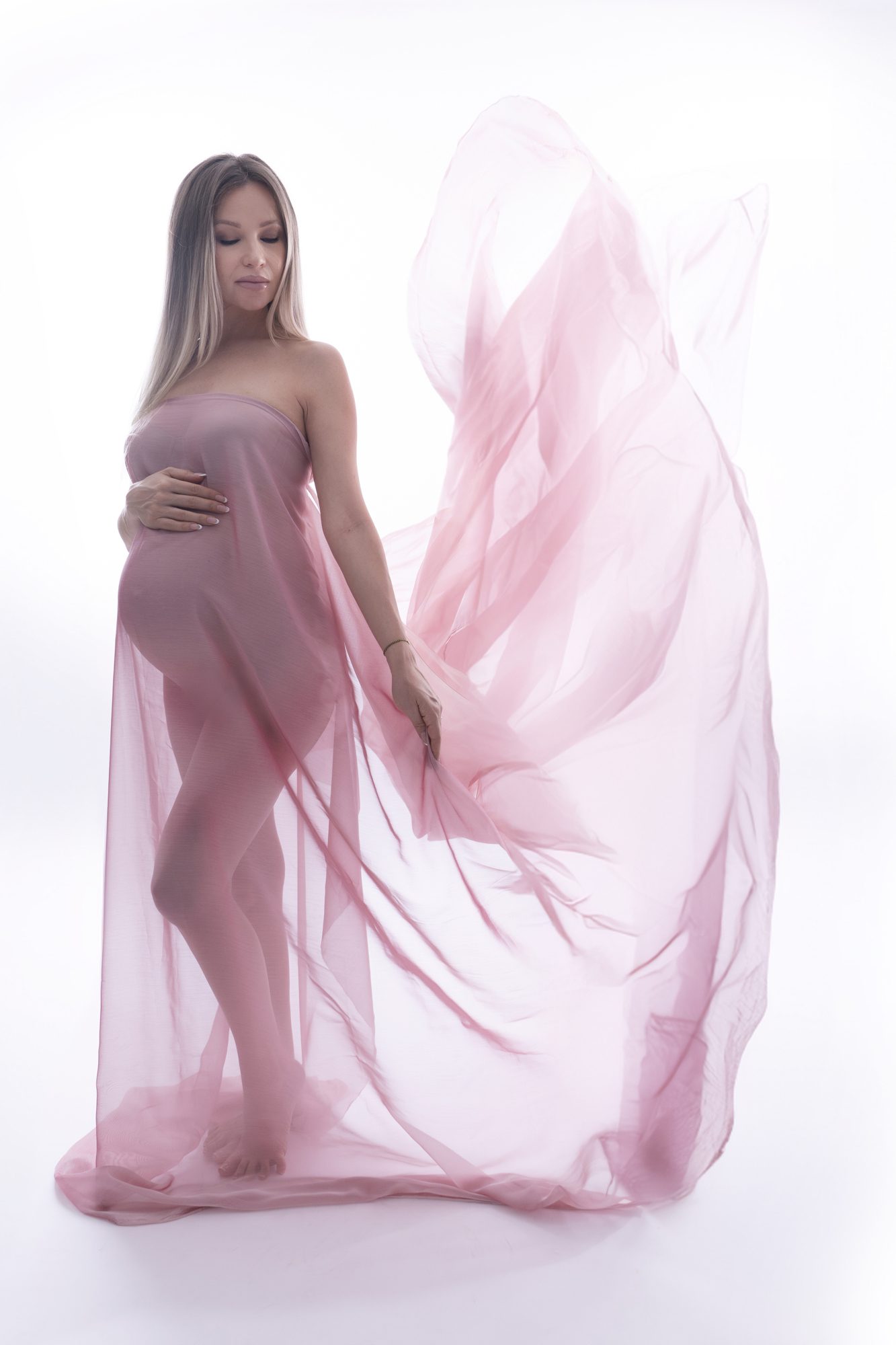 A pregnant woman stands barefoot, draped in flowing pink fabric against a white background, holding her belly with one hand and extending the fabric with the other.