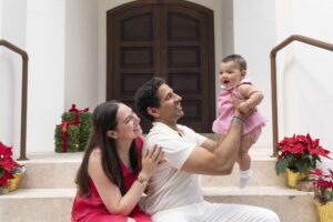 A smiling couple sits on steps holding a baby in pink. Wrapped gifts and poinsettias are beside them, against a large wooden door—capturing a warm holiday moment by a Miami family photographer.