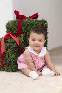 A baby in a red and white outfit sits on a light-colored floor next to two box-shaped green decorations with red ribbons and berries, captured perfectly by a Miami family photographer.