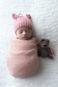 Miami newborn photographer. Newborn baby swaddled in a pink blanket, wearing a pink knit hat with bear ears, lying on a white textured surface next to a small knitted teddy bear.