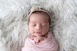 Miami newborn photographer. A newborn baby wrapped in a pink blanket lies on a fluffy white surface, wearing a pink headband with small flowers, eyes closed and hands gently resting near the face.