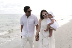 A man, woman, and baby dressed in white walk along a sandy beach near the ocean, holding hands. Captured by a Miami family photographer, the woman holds the baby as they all appear relaxed and happy.