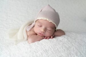 Newborn baby wearing a soft pink hat sleeps on a white textured blanket, resting with head on folded arms.