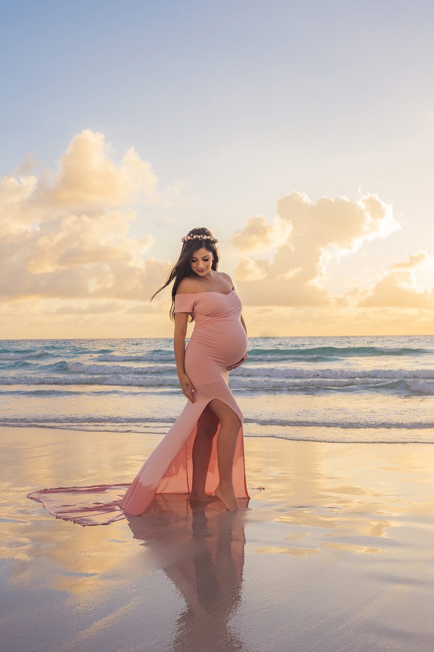 Pregnant woman in a pink dress stands on the beach at sunset, looking down and touching her belly, with waves and clouds in the background.