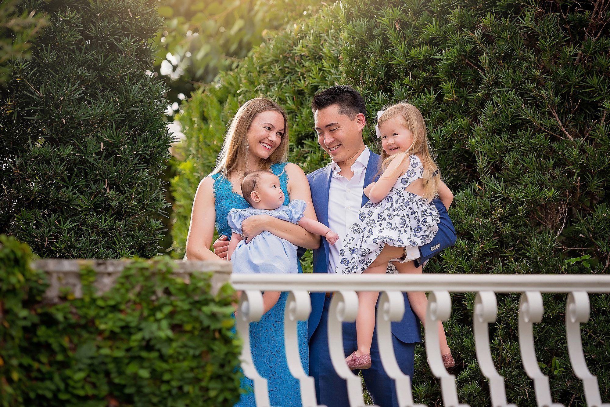 A family of four, with two parents and two young children, stand together outdoors in front of green foliage, smiling and looking at each other.