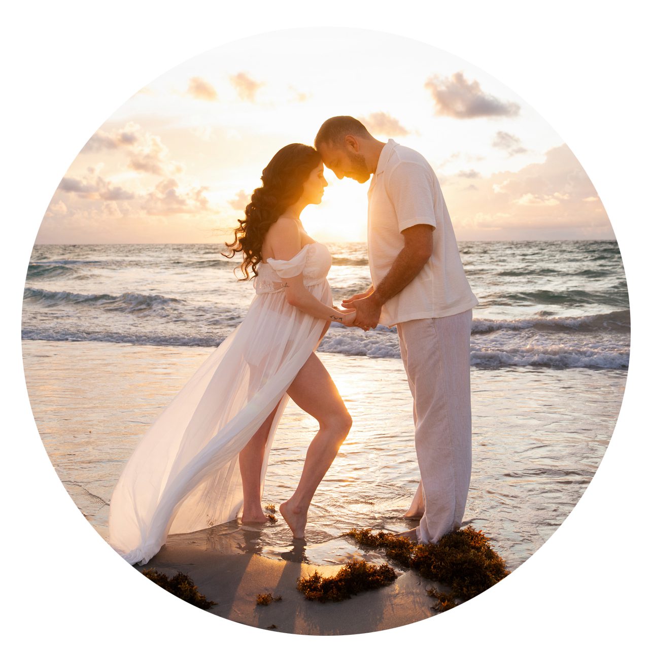 A couple stands barefoot on the beach at sunset, holding hands and facing each other, with the woman's white dress flowing in the breeze and waves in the background.