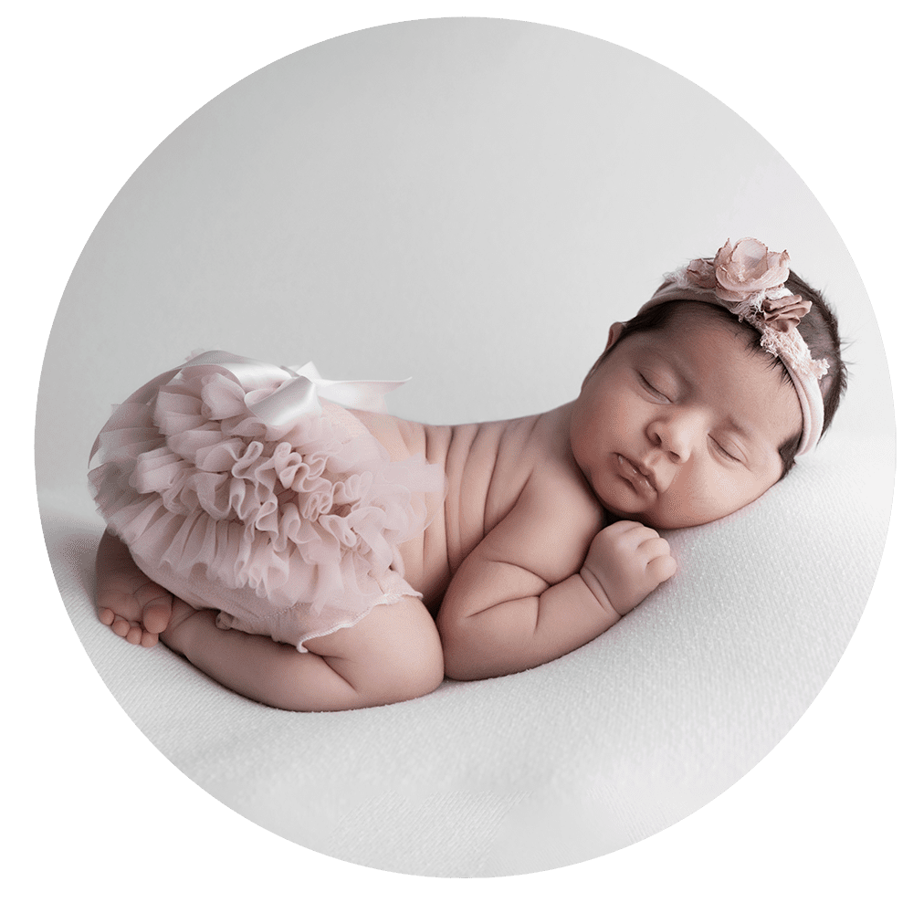 Newborn baby sleeping on a white surface, wearing a pink ruffled skirt and a matching floral headband.