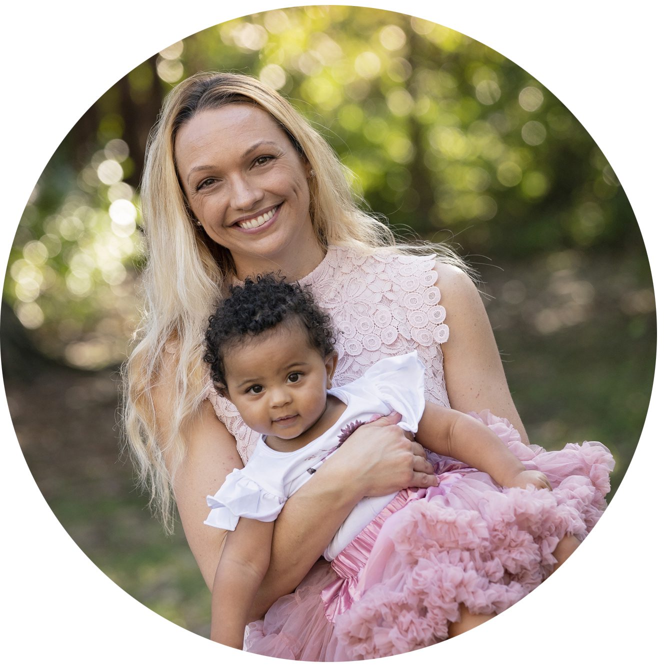 A woman with long blonde hair smiles while holding a young child in a pink dress. They are outdoors with greenery in the background.