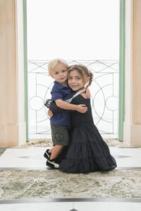 A young boy and girl stand hugging in front of a bright balcony with a metal railing, both dressed in dark blue outfits and smiling at the camera.
