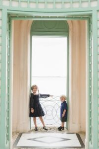 Two children stand in a large arched doorway with a metal railing, overlooking a bright outdoor scene. The girl has braids and wears a dark dress; the younger child also wears dark clothing.