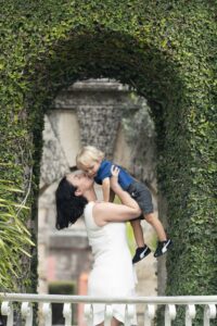 A woman in a white dress lifts a young boy in a blue shirt and gray shorts, kissing his cheek, under an ivy-covered archway.