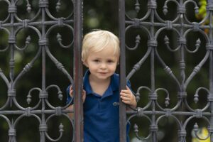 A young child with blond hair and a blue shirt stands behind an ornate metal gate, looking through the bars and smiling slightly.