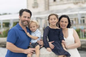 A man, woman, young girl, and young boy pose together outdoors, smiling at the camera with a building and greenery in the background.