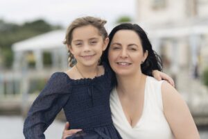 A woman and a young girl with braided hair stand close together outdoors, both smiling at the camera.