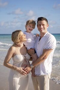 A man, woman, and young boy pose together on a sunny beach during a Miami Beach maternity family photoshoot. The smiling, pregnant woman looks at the child in the man's arms, with the ocean and sky behind them.