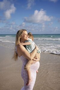 A woman in a white dress stands on a beach, holding a young child in her arms with waves and a partly cloudy sky behind her during a Miami Beach maternity family photoshoot.