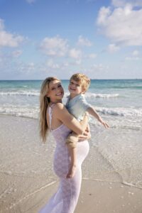 A woman in a white dress holds a smiling young boy on the sandy shore during a Miami Beach maternity family photoshoot, with gentle waves and a bright blue sky in the background.