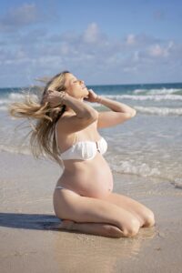 A pregnant woman in a white bikini kneels on the sandy shore, eyes closed, with hands in her hair, near gentle ocean waves under a partly cloudy sky during a Miami Beach maternity family photoshoot.