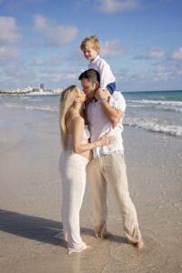 A man holds a young boy on his shoulders while standing on the beach with a woman during their Miami Beach maternity family photoshoot. All three are dressed in light hues, with the ocean and sky providing a scenic backdrop.