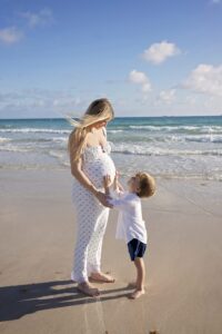 A pregnant woman stands on the sand during a Miami Beach maternity family photoshoot, while a young boy touches her belly. The ocean waves and blue sky are visible in the background.