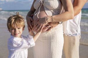 A pregnant woman stands on the beach with a man and a young boy, all touching her belly during their Miami Beach maternity family photoshoot. The boy is smiling at the camera.