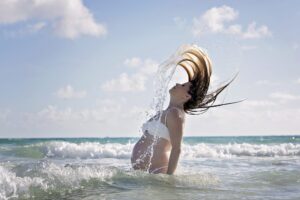 A pregnant woman in a white bikini flips her wet hair back while standing in ocean waves under a partly cloudy sky during a Miami Beach maternity family photoshoot.