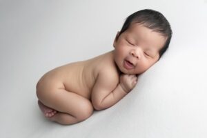 A newborn baby is lying on their side on a white blanket, eyes closed, with a relaxed and peaceful expression.