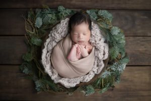 Newborn baby wrapped in a beige blanket, sleeping in a nest-like arrangement with green fir branches on a wooden surface.