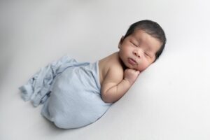 Newborn baby sleeping on a white surface, wrapped in a light blue blanket, lying on their side with one hand under their cheek—a serene moment captured by Miami newborn photography.