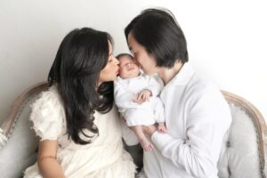 A couple sits on a sofa, each gently kissing their newborn who is wrapped in a white blanket and cradled in their arms.