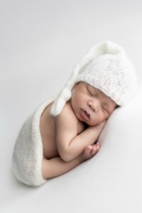 Newborn baby sleeping on side, wearing a white knit hat and wrap, on a soft white surface—capturing the pure innocence of Miami newborn photography.