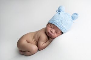A newborn baby wearing a light blue knit hat with bear ears sleeps on a white surface, lying on their side with legs curled up—a sweet moment captured by Miami newborn photography.