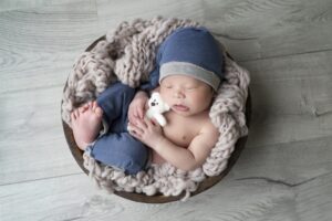 Newborn baby wearing a blue hat and pants sleeps in a round wooden bowl with a chunky knit blanket, holding a small white stuffed bear.