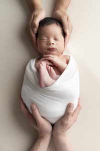 A newborn baby wrapped in a white blanket is cradled gently by adult hands, resting on a light-colored surface.
